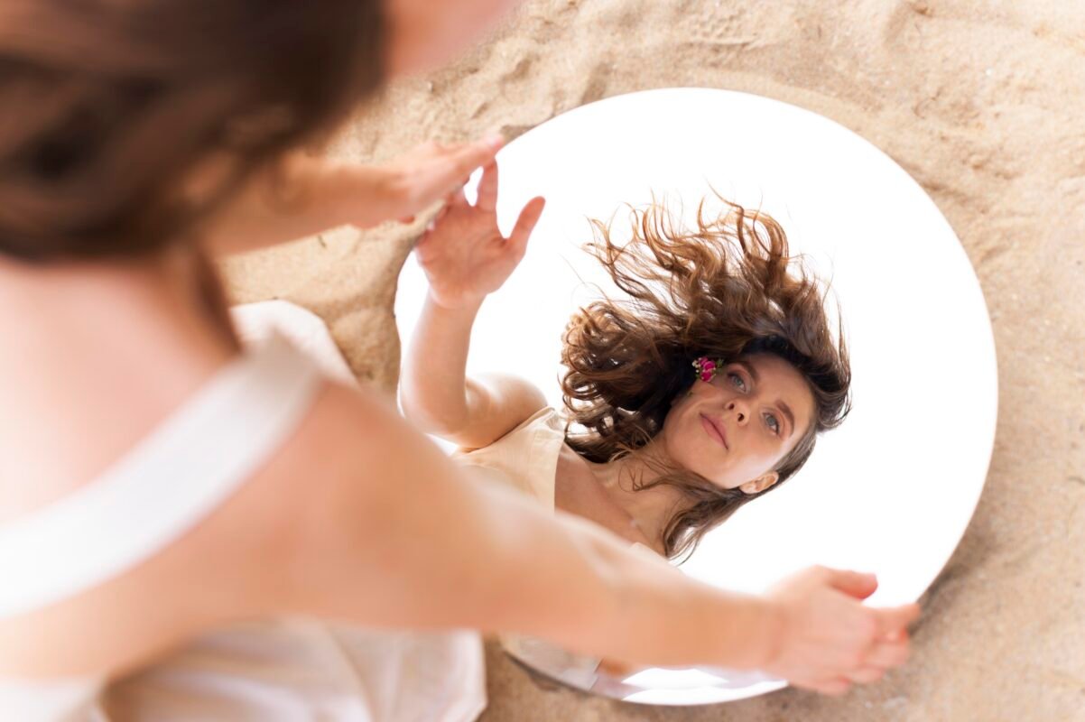 This image features a woman standing outdoors, posing with confidence while holding a round mirror. The scene symbolises self-love, empowerment, and embracing the best version of oneself. The natural setting enhances the feeling of authenticity and personal strength